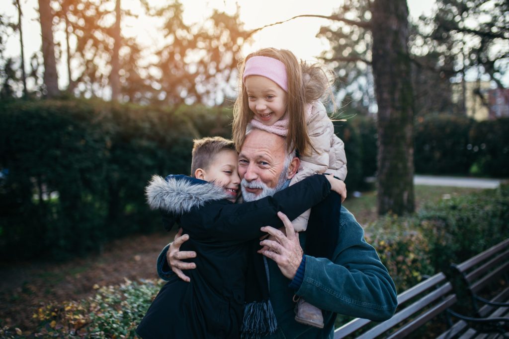 Elderly man in a park hugging his two grandchildren next to a bench.